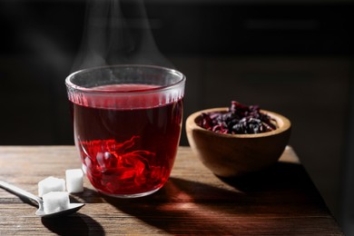 Delicious hibiscus tea in glass, sugar cubes and dry roselle petals on wooden table, closeup