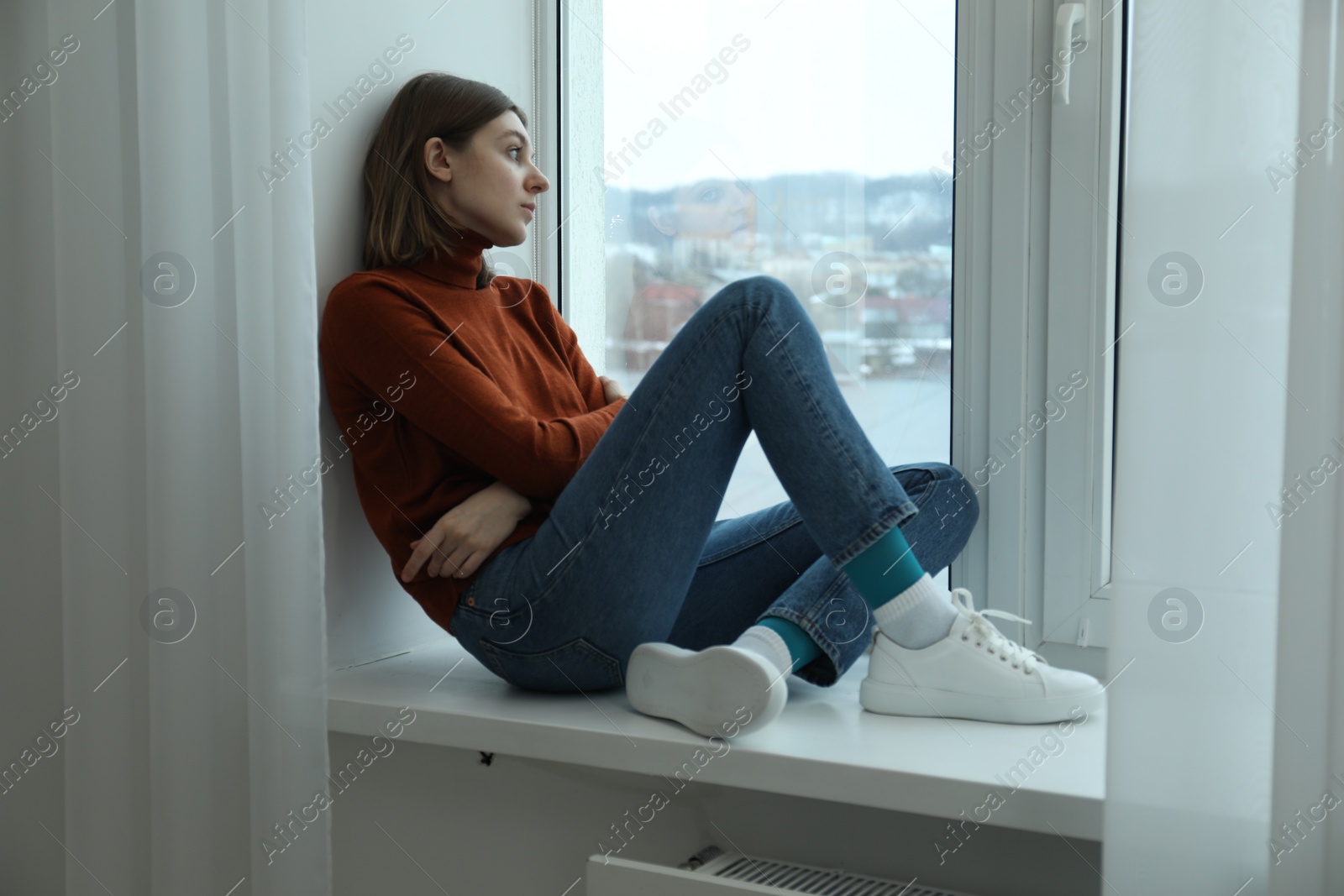 Photo of Sad young woman sitting on windowsill near window at home