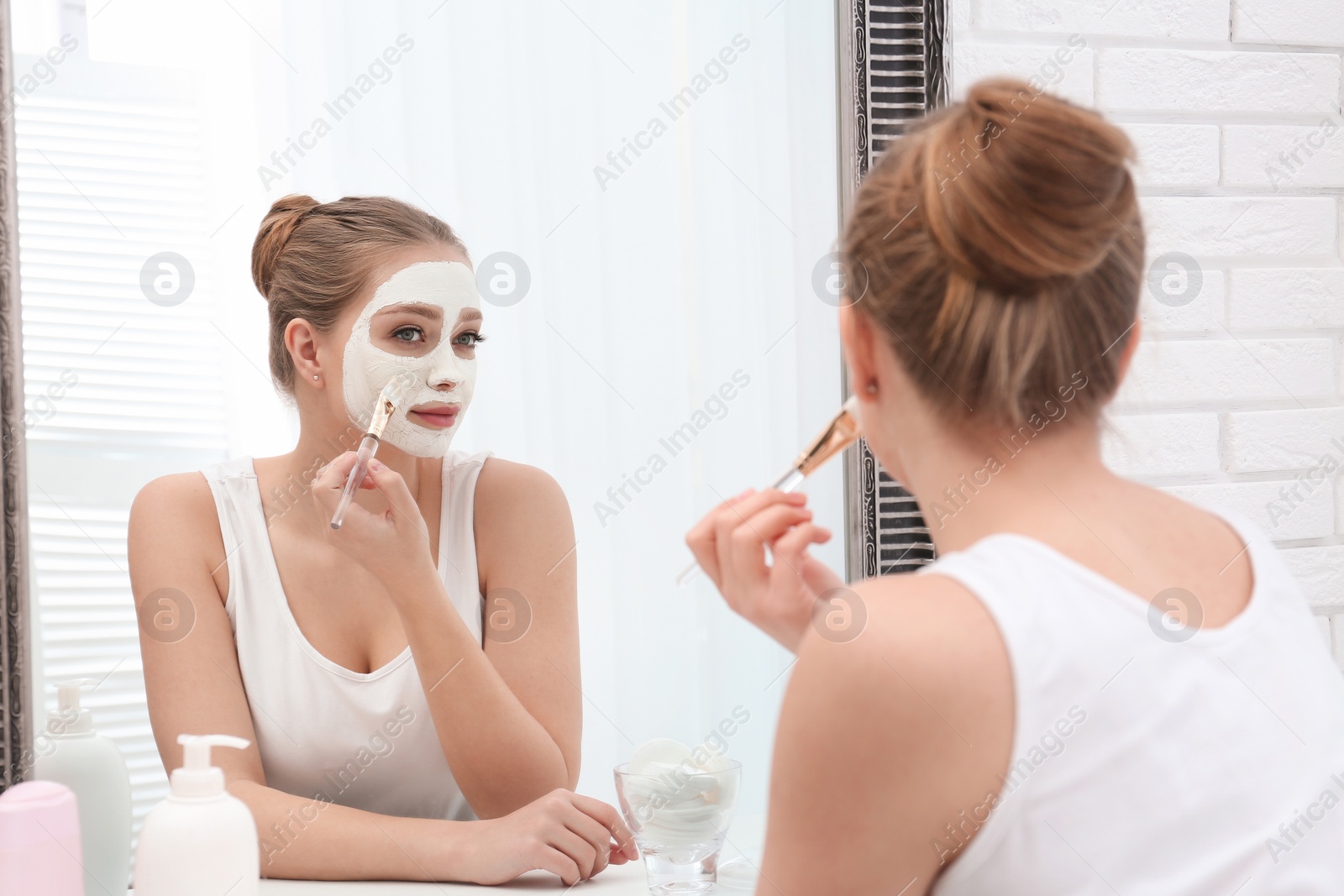 Photo of Beautiful woman applying homemade clay mask on her face at mirror indoors