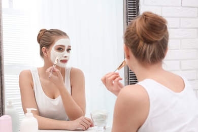 Beautiful woman applying homemade clay mask on her face at mirror indoors