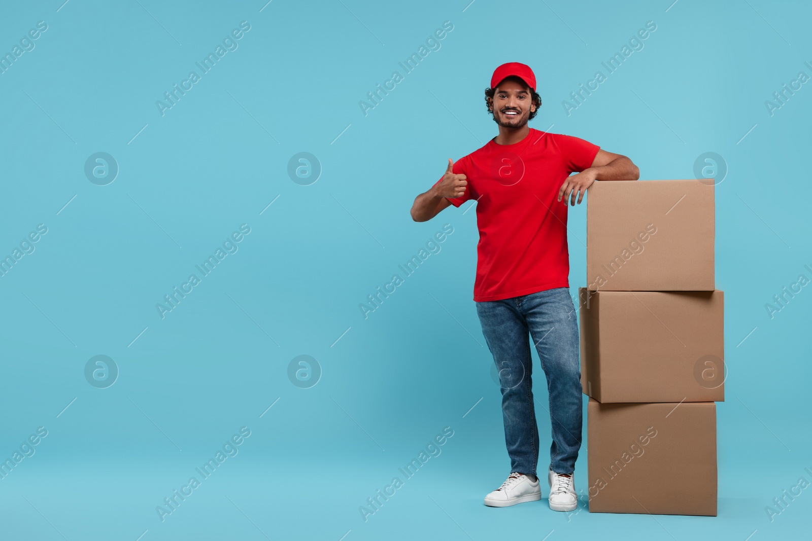Photo of Happy courier with stack of parcels showing thumb up on light blue background, space for text