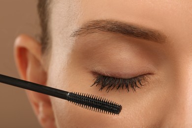 Woman applying mascara onto eyelashes against light brown background, closeup