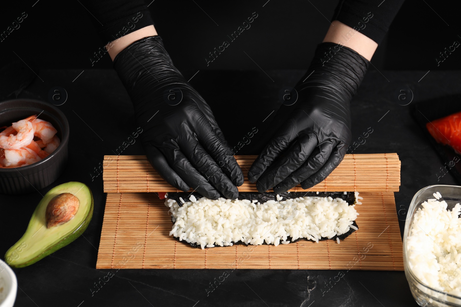 Photo of Chef in gloves wrapping sushi roll at dark textured table, closeup