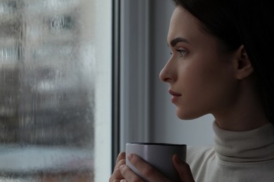 Photo of Melancholic young woman with cup of drink near window on rainy day, space for text. Loneliness concept