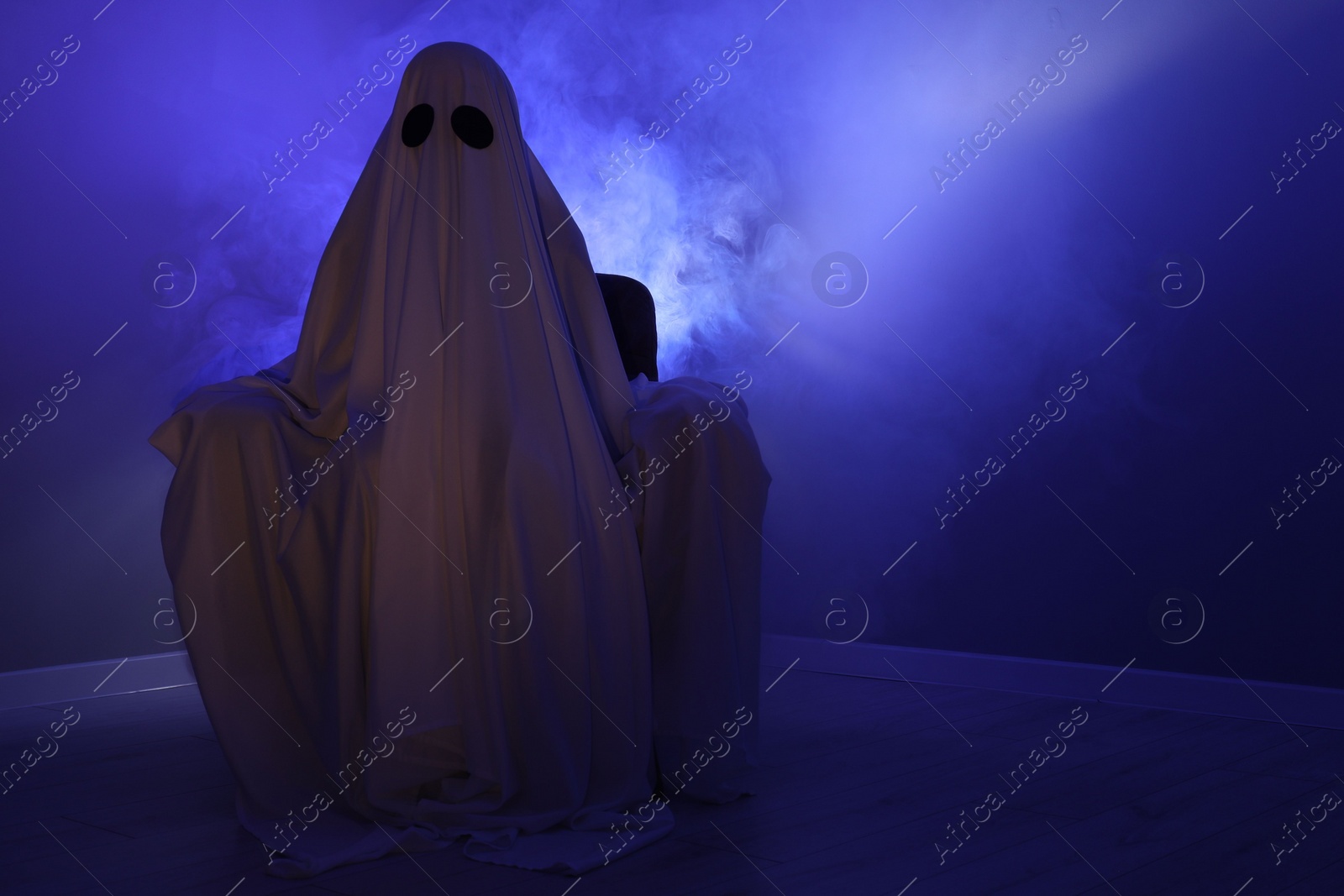 Photo of Creepy ghost. Woman covered with sheet sitting in armchair in blue light, space for text