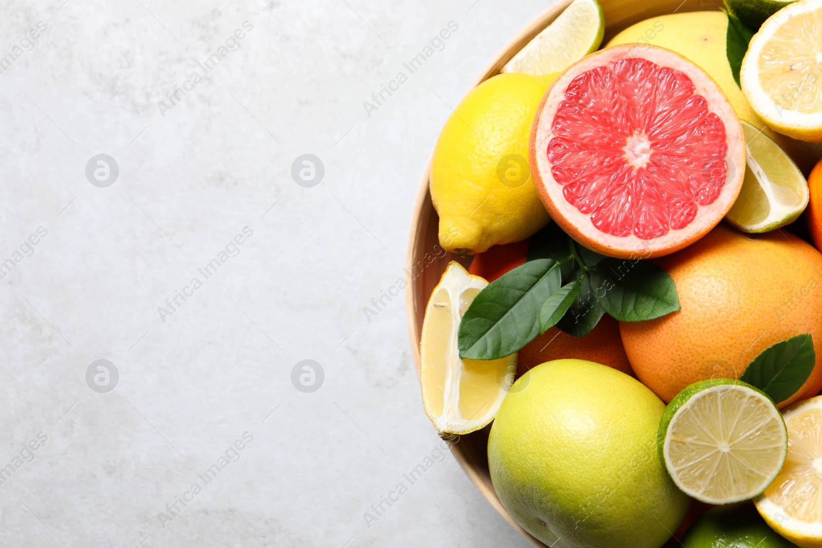 Photo of Different fresh citrus fruits and leaves in bowl on light table, top view. Space for text