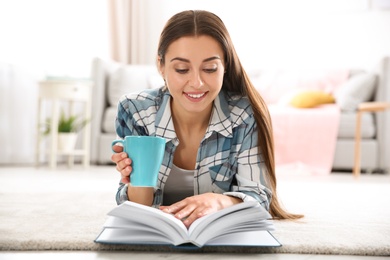 Photo of Beautiful young woman with cup of coffee reading book on floor at home