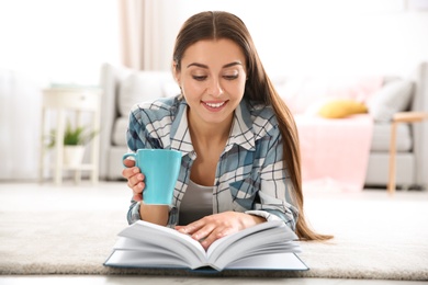 Beautiful young woman with cup of coffee reading book on floor at home