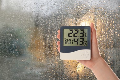 Image of Woman holding digital hygrometer with thermometer near window on rainy day, closeup. Space for text