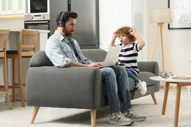 Photo of Little boy bothering his father at home. Man with laptop and headphones working remotely