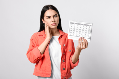 Photo of Pensive young woman holding calendar with marked menstrual cycle days on light background