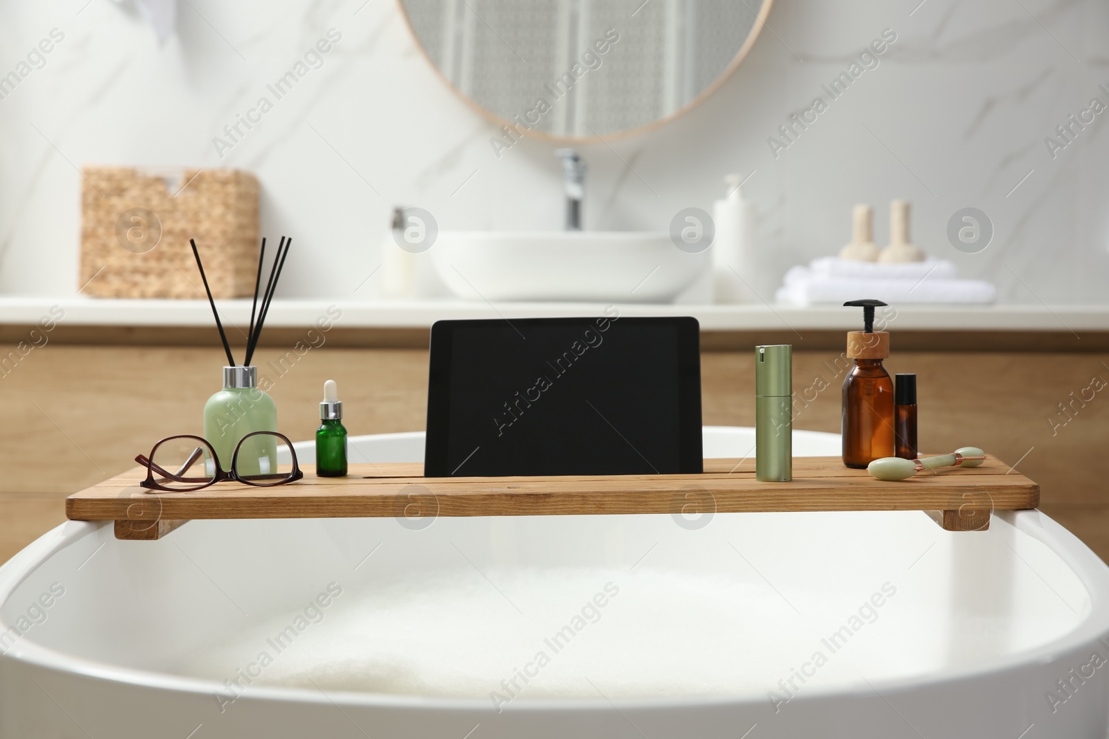 Photo of Wooden bath tray with tablet, face roller and cosmetic products on tub indoors