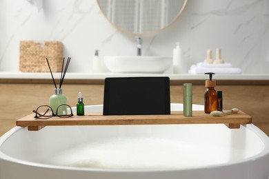 Photo of Wooden bath tray with tablet, face roller and cosmetic products on tub indoors