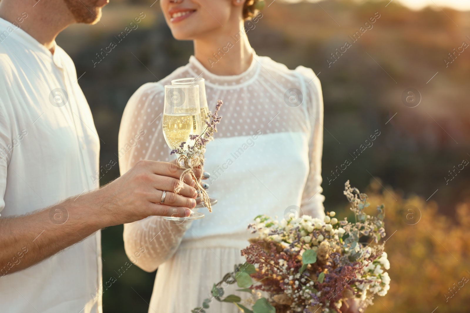 Photo of Happy newlyweds with beautiful field bouquet and glasses of champagne outdoors, closeup