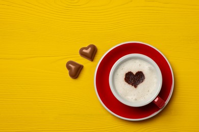 Photo of Cup of aromatic coffee with heart shaped decoration and chocolate candies on yellow wooden table, flat lay. Space for text