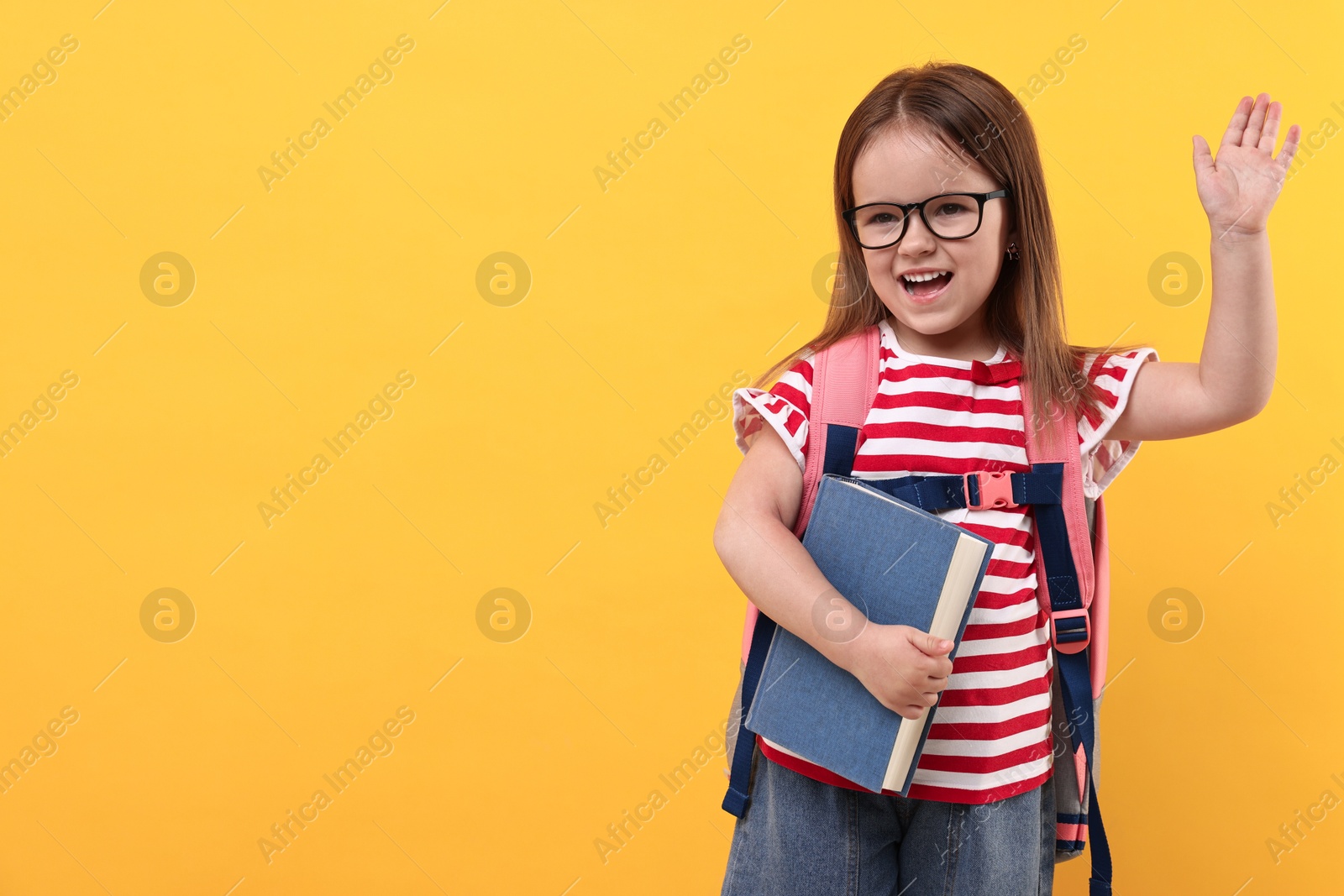 Photo of Cute little girl with book and backpack greeting someone on orange background. Space for text