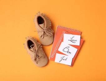 Notebooks with different baby names and child's shoes on orange background, flat lay