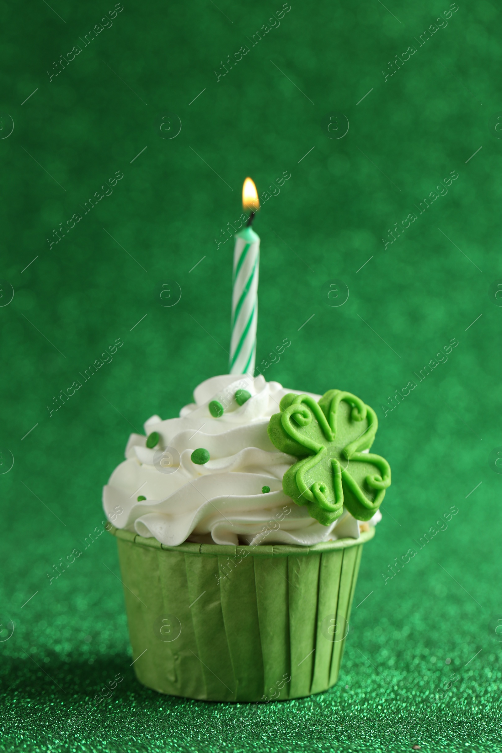 Photo of St. Patrick's day party. Tasty cupcake with clover leaf topper and burning candle on shiny green surface, closeup