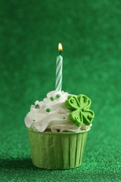 St. Patrick's day party. Tasty cupcake with clover leaf topper and burning candle on shiny green surface, closeup