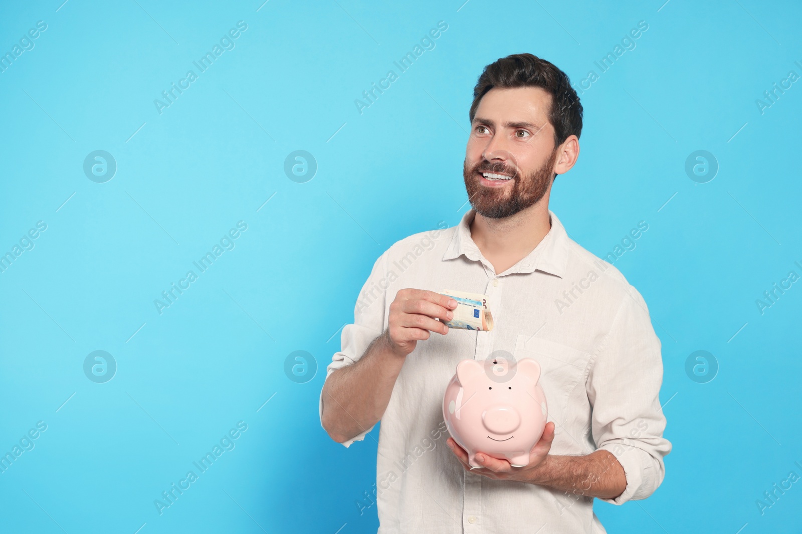 Photo of Happy man putting money into piggy bank on light blue background, space for text