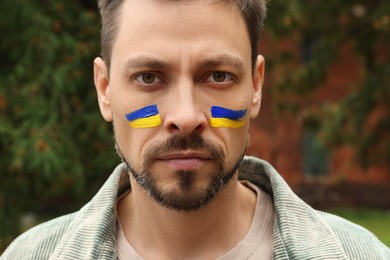 Photo of Angry man with drawings of Ukrainian flag on face outdoors, closeup