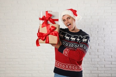 Happy man in Christmas sweater and Santa hat holding gift boxes near white brick wall