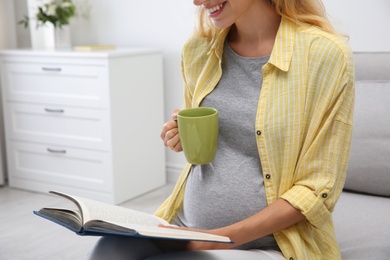 Photo of Beautiful pregnant woman with book drinking tea at home, closeup