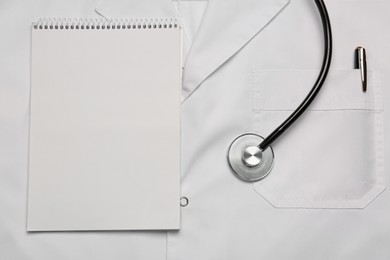 Stethoscope and notepad on white medical uniform, top view