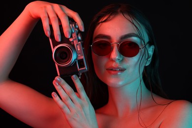 Beautiful woman with sunglasses and vintage camera posing in neon lights against black background, closeup