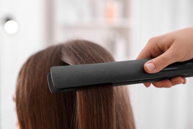 Photo of Hairdresser straightening woman's hair with flat iron indoors, closeup