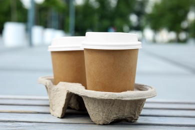 Photo of Takeaway paper coffee cups with plastic lids in cardboard holder on wooden bench outdoors