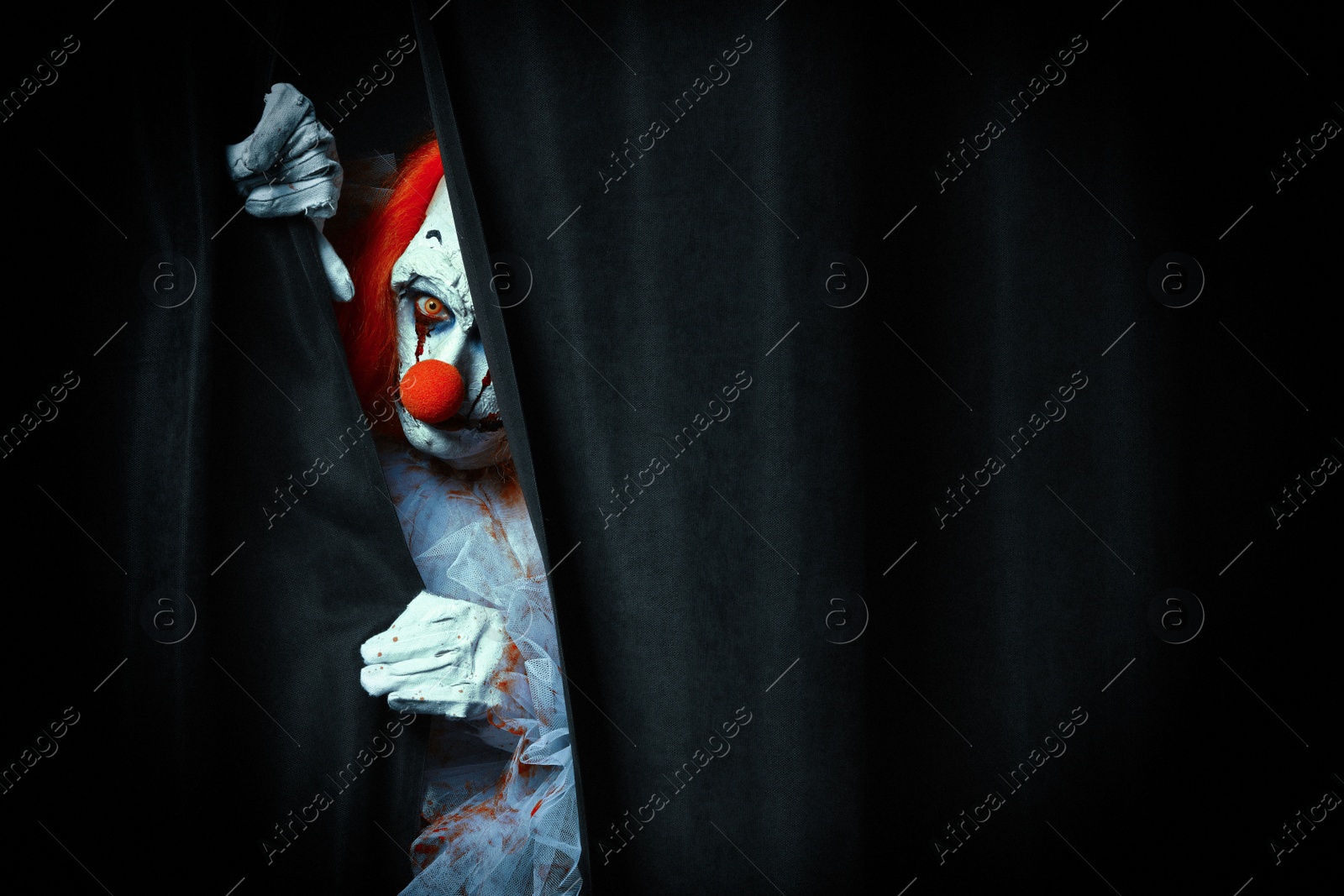 Photo of Terrifying clown hiding behind black curtains, space for text. Halloween party costume