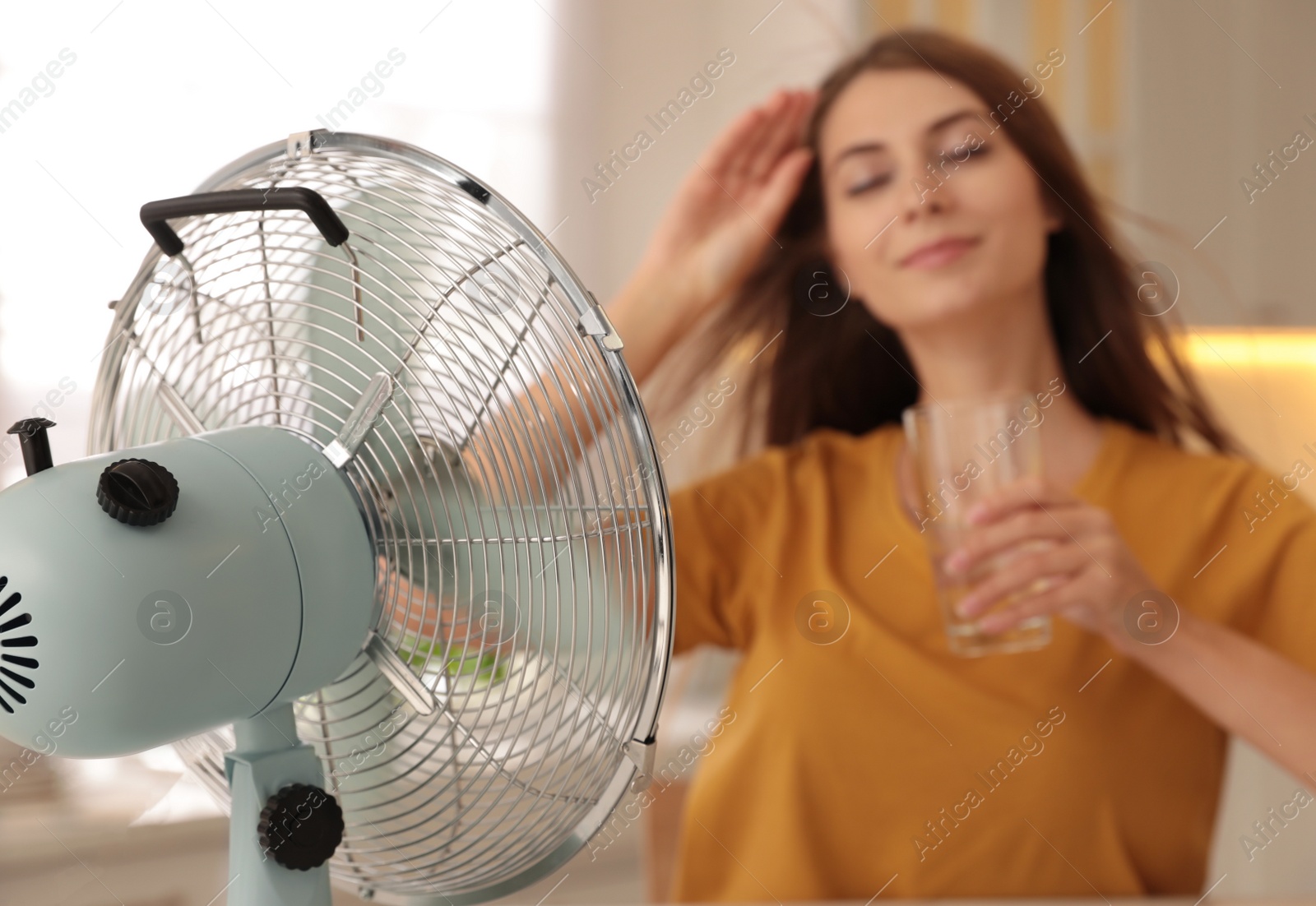 Photo of Woman with glass of water in kitchen, focus on fan. Summer heat