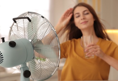 Photo of Woman with glass of water in kitchen, focus on fan. Summer heat