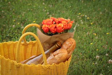 Photo of Yellow wicker bag with beautiful roses, bottle of wine and baguettes on green grass outdoors