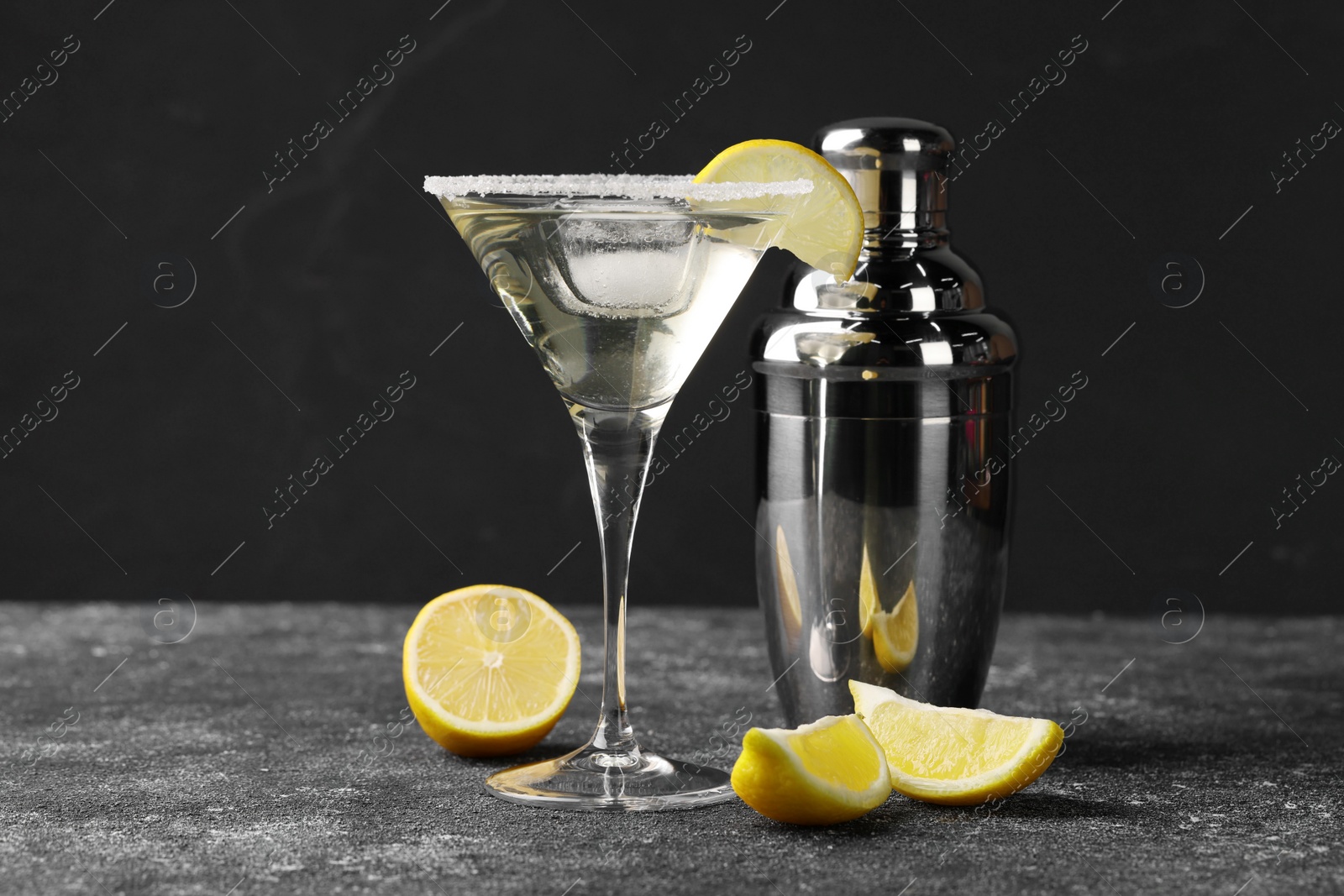 Photo of Martini glass of refreshing cocktail with lemon and ice cube near shaker on black textured table