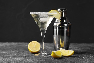 Photo of Martini glass of refreshing cocktail with lemon and ice cube near shaker on black textured table