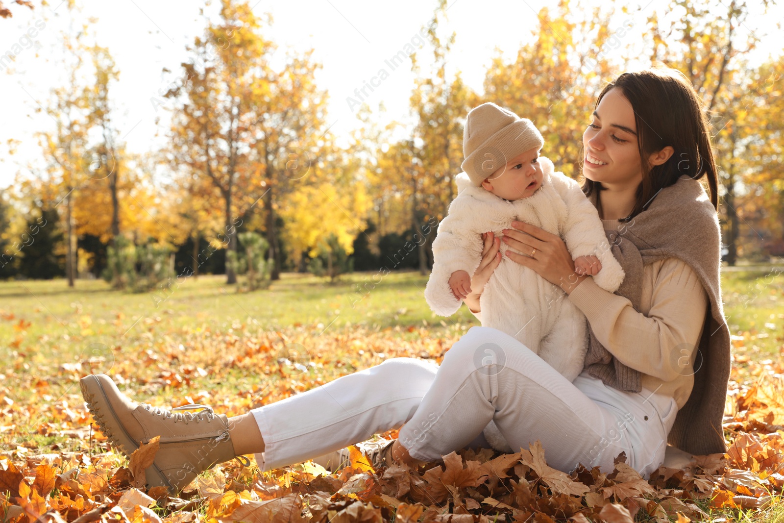 Photo of Happy mother with her baby son sitting on fallen leaves in autumn park, space for text