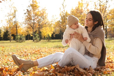 Happy mother with her baby son sitting on fallen leaves in autumn park, space for text