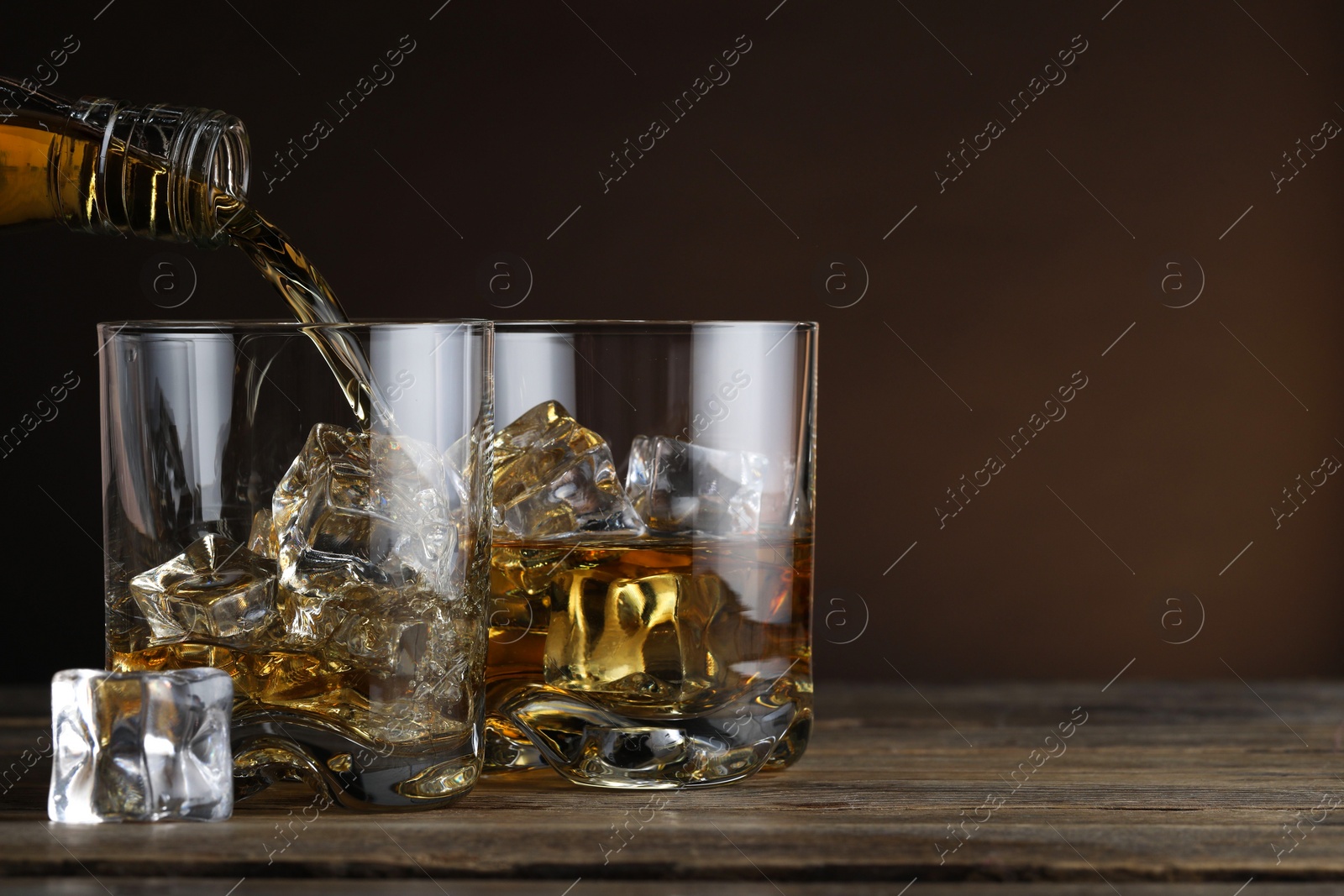 Photo of Pouring whiskey into glass with ice cubes at wooden table against brown background, space for text