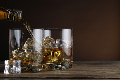Photo of Pouring whiskey into glass with ice cubes at wooden table against brown background, space for text