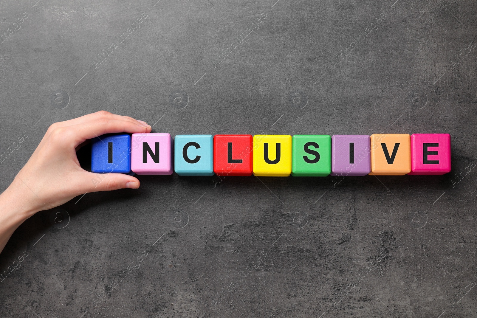 Photo of Woman making word Inclusive with cubes on dark grey background, top view