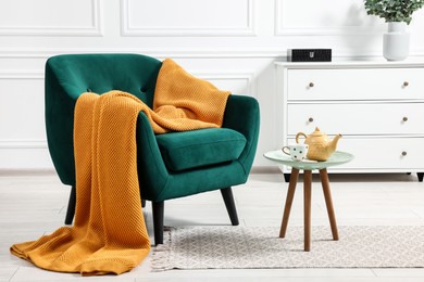 Comfortable armchair with blanket, side table and chest of drawers in room