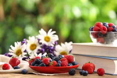 Photo of Different fresh ripe berries, beautiful flowers and books on wooden table outdoors