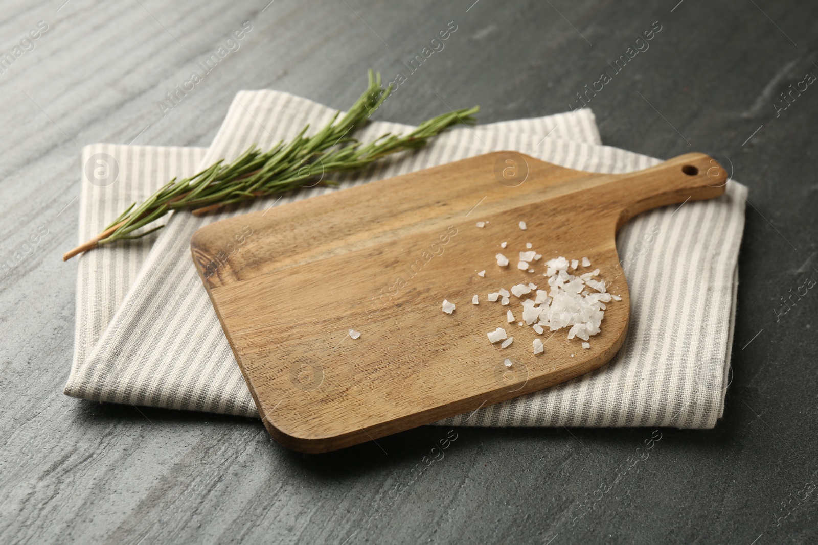 Photo of Wooden cutting board, kitchen towel, rosemary and salt on dark grey table