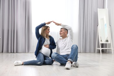 Photo of Young family housing concept. Pregnant woman with her husband forming roof with their hands while sitting on floor at home