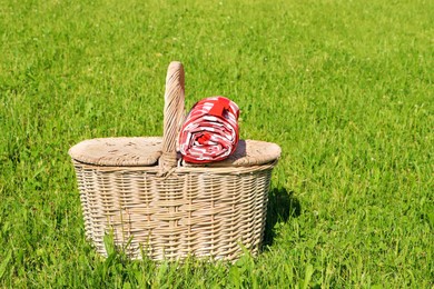 Photo of Rolled checkered tablecloth with picnic basket on green grass outdoors, space for text