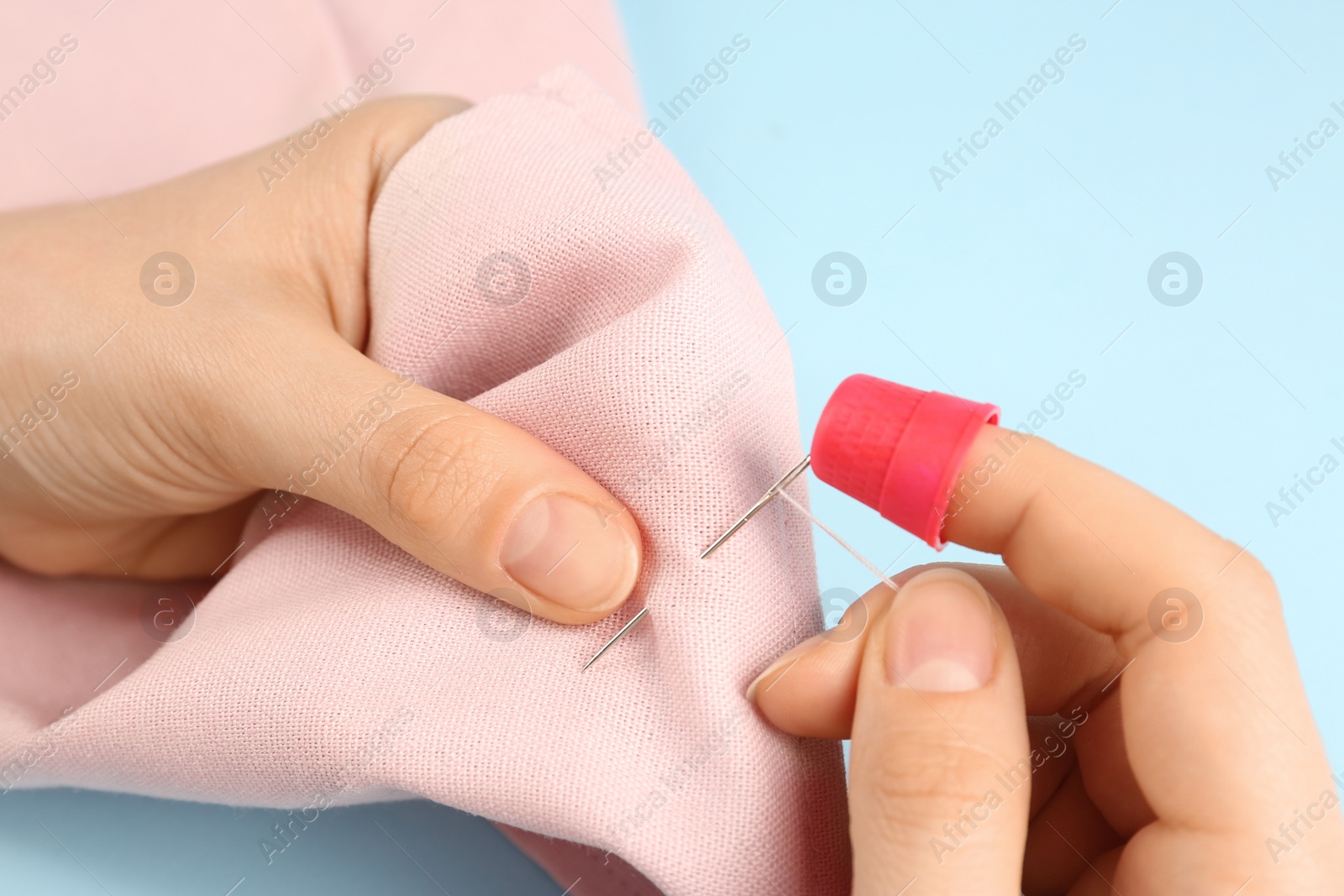 Photo of Woman sewing on pink fabric with thimble and needle against light blue background, closeup