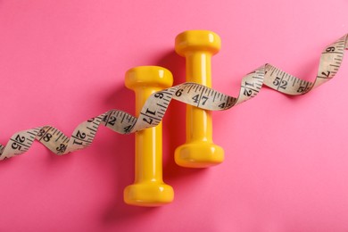 Measuring tape and dumbbells on pink background, flat lay. Weight control concept