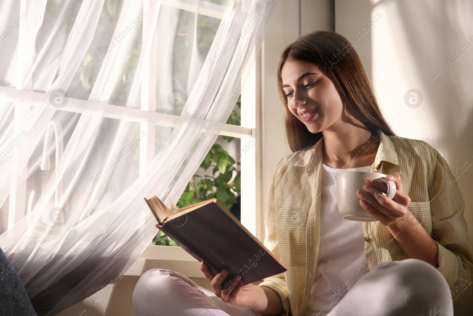 Photo of Beautiful young woman reading book near window at home